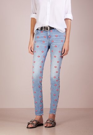 Slim-fit lyseblå jeans med røde hjerte-mønstre og tekst, kombineret med en hvid skjorte, sort bælte og sandaler med metal spænde.