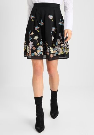 Czarna plisowana spódnica z kolorowymi haftowanymi ptakami i kwiatami, noszona z czarnymi botkami i białą bluzką z długim rękawem.