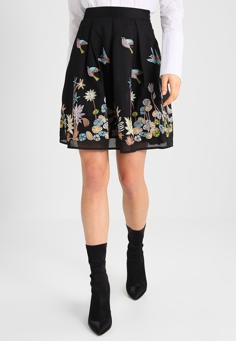 Jupe plissée noire ornée de broderies colorées d'oiseaux et de fleurs, portée avec des bottines noires et un haut blanc à manches longues.