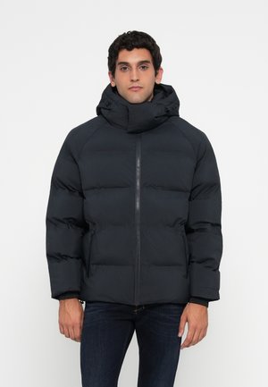 Hombre vestido con una chaqueta acolchada negra de cuello alto y cremallera frontal, de pie frente a un fondo blanco liso.