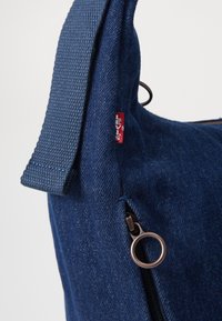 Sac en denim bleu foncé avec un tissu texturé, doté d'une fermeture zippée et d'un tirette en bronze. Sangle en tissu ajustable visible.