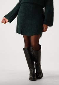 Kvinne iført en mørkegrønn, fluffy strikket skjørt og matchende genser, gjennomsiktige sorte strømpebukser og høye, sorte lærstøvler på hvit bakgrunn.