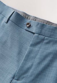 Pantalon de costume bleu clair avec fermeture par bouton et passants de ceinture, montrant la doublure intérieure grise à motifs de la ceinture.