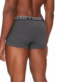 Boxers en coton gris avec une coupe ajustée, présentant une ceinture noire portant l'inscription "GUESS" en lettres blanches.