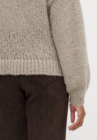 Vaaleanbeige neulottu villapaita, jossa on väljä istuvuus ja tekstuurinen kuvio, ribbed hihansuut ja helma, yhdistettynä tummanruskeisiin housuihin.