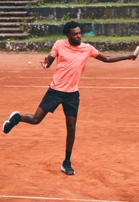 Idrettsutøver i en lys korallfarget skjorte og svarte shorts, midt i en handling på en leirgulv tennisbane, iført svarte joggesko med hvite såler.