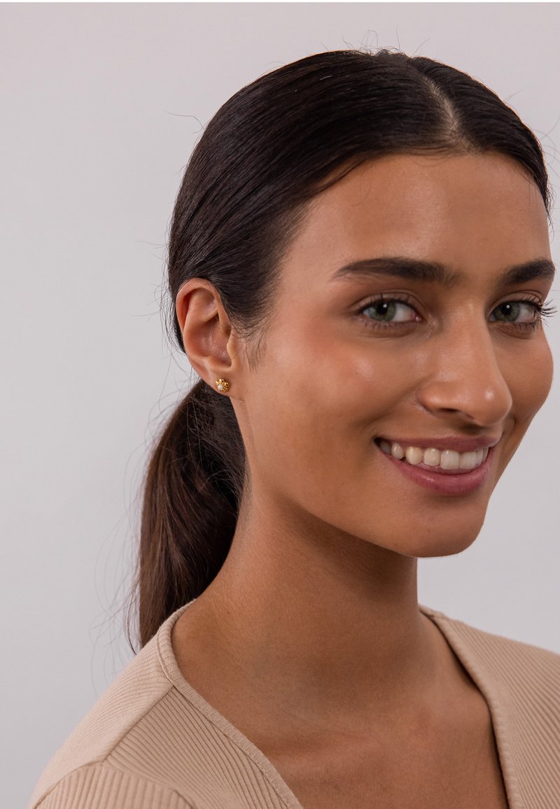 Goldene Steckerohrringe mit floralem Design, die kleine Blütenblätter und eine strukturierte Oberfläche aufweisen, getragen von einer Person mit dunklen Haaren und einem neutralen Oberteil.