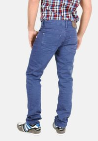 Los jeans de algodón azul presentan un corte ajustado, bolsillos traseros y una textura sutil. Combinados con zapatillas deportivas negras y azules.