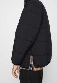 Svart vadderad jacka med quiltad design, utrustad med hög krage och dragkedja. Sedd från sidan, vilket visar en lager-på-lager-look.