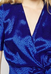 Tissu texturé bleu royal avec un motif animalier discret. Conception en V avec des manches courtes et un détail de taille froncée.