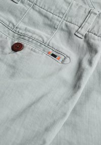 Lichtgrijze broek van zacht materiaal met een bruine knoop, voorzien van een kleurrijke stikselafwerking in oranje en zwart op de achterzak.