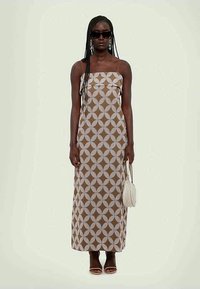 Maksivakars ar ģeometrisku brūnu un baltu rakstu, bez piedurknēm un taisnu piegriezumu. Aksesuāri: saulesbrilles un maza soma.