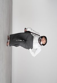 Žena u bijeloj bluzici, sivoj širokoj hlačama, japankama, šarenim maramom oko vrata i drži smeđu torbu preko ramena na neutralnoj pozadini.