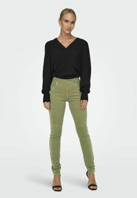 Blouse noire à manches longues avec un décolleté en V, associée à un pantalon skinny vert clair. Des sandales à talons noires complètent la tenue, offrant un look élégant.