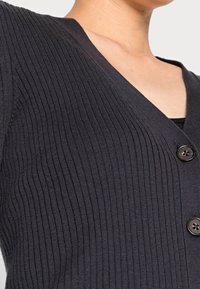 Cardigan a coste scura con scollatura a V e due bottoni frontali. Il materiale sembra morbido con una finitura testurizzata.