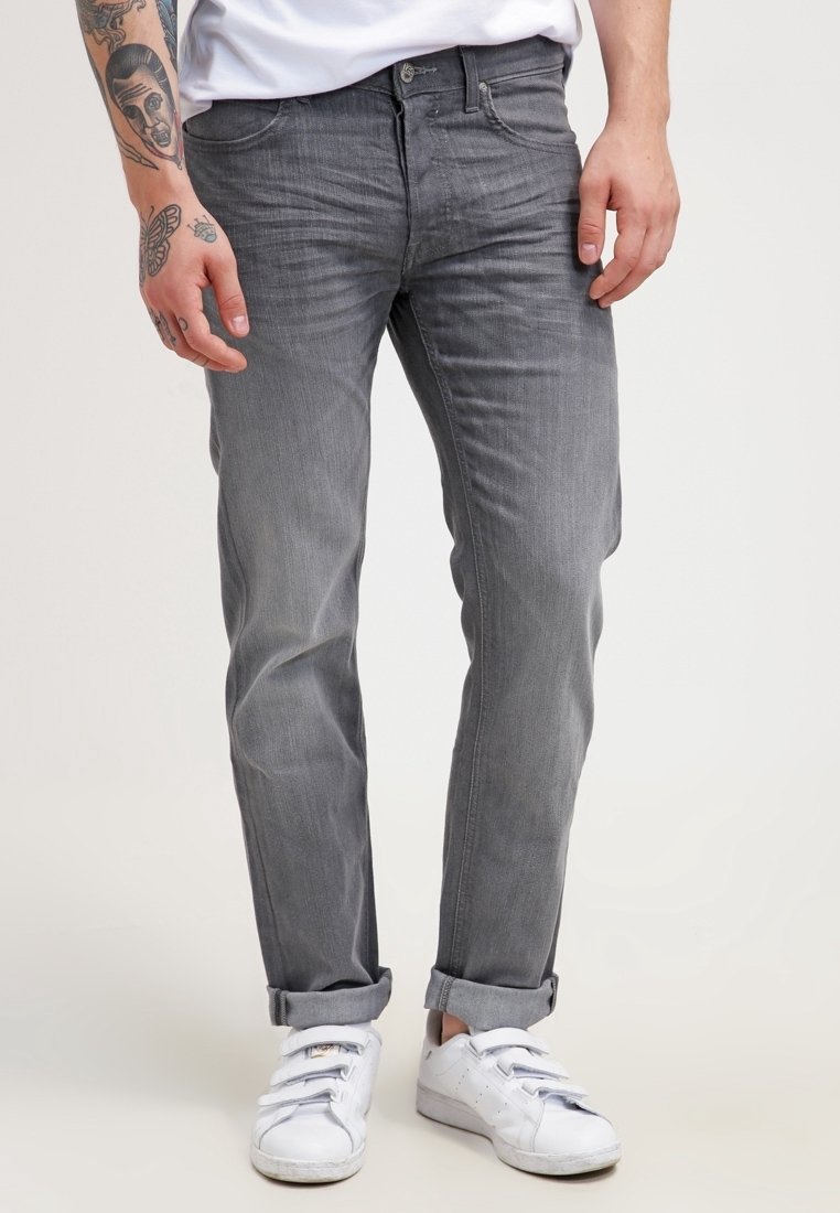 Homme en jean gris retroussé, baskets blanches et chemise blanche, montrant des tatouages sur l'avant-bras gauche et la main sur un fond uni.