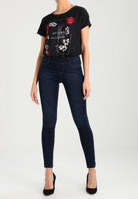 Top negro de manga corta con estampado floral y el texto "FLORES DE OTOÑO", combinado con jeans ajustados de mezclilla oscura y tacones altos negros.