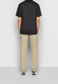 Černé tričko s krátkým rukávem, béžové cargo kalhoty s bočními kapsami, černo-bílé tenisky. Jednoduché pozadí, důraz na detaily oblečení.