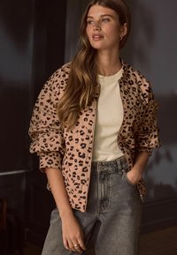 Leopardmønstret bomberjakke i lys beige, med ribbede manchetter og lynlås. Båret over en cremefarvet tætsiddende top og parret med højtaljede jeans.