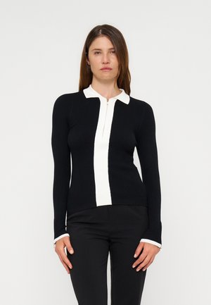 Femme aux cheveux bruns et raides porte un haut noir à manches longues avec une bande centrale blanche, col et poignets, associé à un pantalon noir.