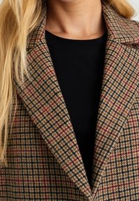 Brun och beige houndstoothmönstrad ullkappa med notskanskrage, bärs över en svart topp med en strukturerad tygfinish.