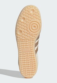 Semelle d'une sneaker avec une base en caoutchouc texturée de couleur crème. Présente des motifs circulaires et hexagonaux, avec un logo en relief centré.