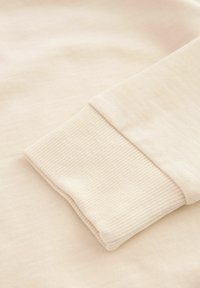 Beige katoenen stof met een ribbelstructuur op de manchetten. Het materiaal lijkt zacht en licht rekbaar, wat het comfort en de pasvorm verbetert.