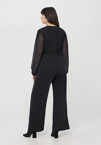 Czarny kombinezon z długimi przezroczystymi rękawami, dopasowanym korpusem i szerokimi nogawkami. Zawiera detal w postaci ściągniętej talii. Noszony z czarnymi szpilkami.