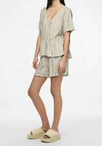 Completo a due pezzi a righe in beige e bianco, con un top a maniche corte e scollo a V, abbinato a pantaloni corti con volant, e sandali slide di colore chiaro.