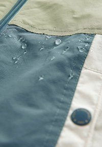 Des gouttes d'eau reposent sur un tissu ripstop bleu et beige avec des coutures visibles et un bouton rond près du bord du tissu.