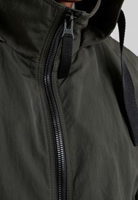 Khaki hooded jacket featuring a zip closure, textured fabric, and a logo tag. It has a smooth finish with adjustable drawstring accents.