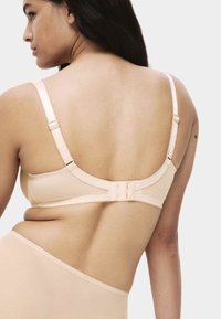 Femme portant un soutien-gorge beige et un bas taille haute assorti, vue de dos sur un fond clair.