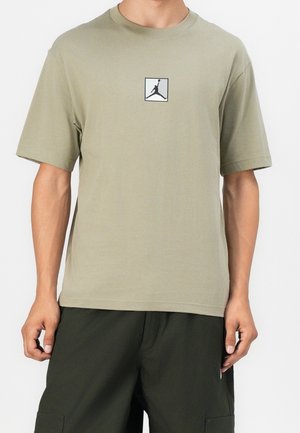 Uomo che indossa una T-shirt manica corta verde oliva con il logo Jumpman di basket in bianco e nero sul petto e pantaloni verde scuro.