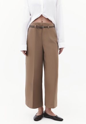 Persoon draagt hoog getailleerde, wijde bruine broek met een smalle bruine riem, een cropped wit shirt met lange mouwen en zwarte platte schoenen.