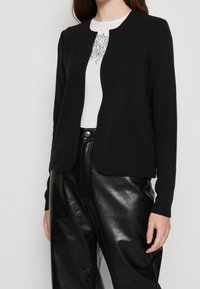 Czarna teksturowana kurtka z okrągłym dekoltem i otwartym przodem, noszona na białym T-shircie i czarnych wysokim stanem skórzanych spodniach.