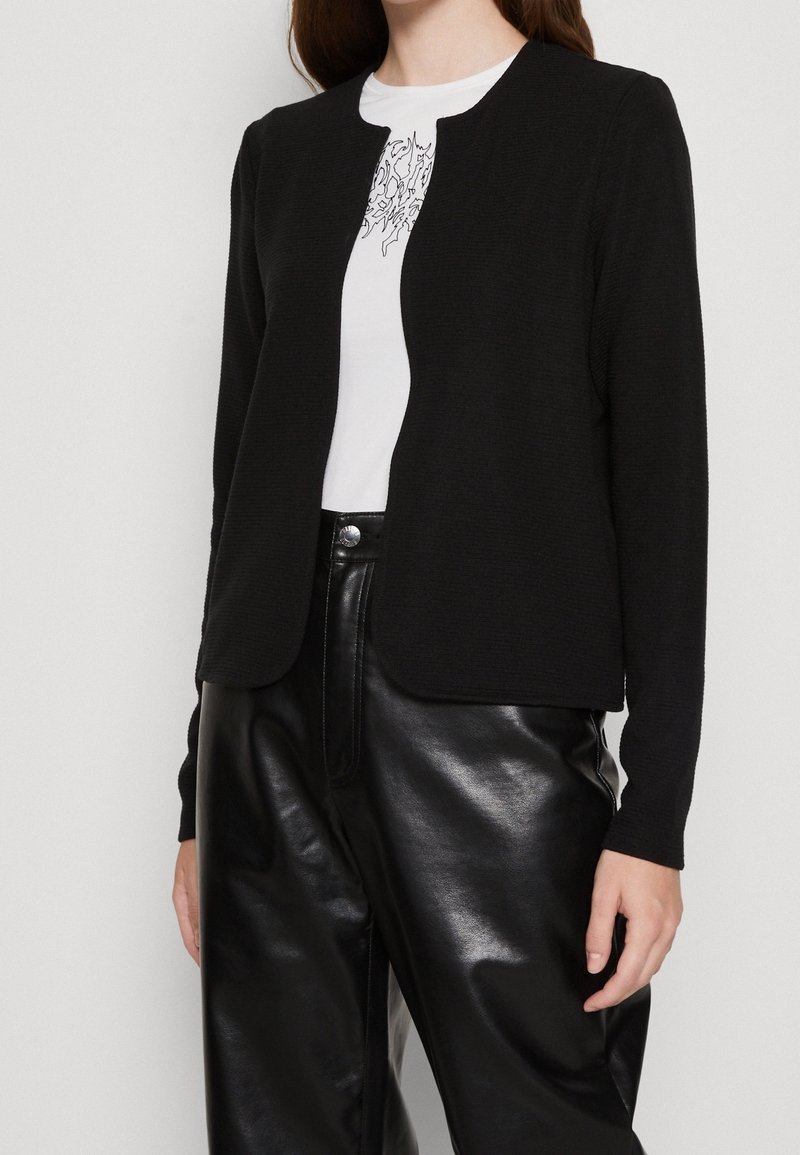 Czarna teksturowana kurtka z okrągłym dekoltem i otwartym przodem, noszona na białym T-shircie i czarnych wysokim stanem skórzanych spodniach.