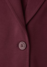 Gros plan sur un blazer en tissu bordeaux avec un seul bouton rond portant l'inscription "Street One" sur le revers avant.