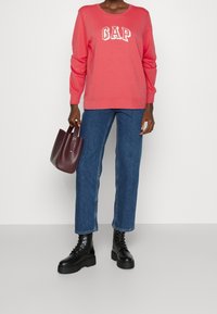Persona vestida con sudadera coral de GAP, pantalones vaqueros azules, botas negras con cordones, sosteniendo un pequeño bolso de cuero marrón oscuro, de pie contra una pared lisa.