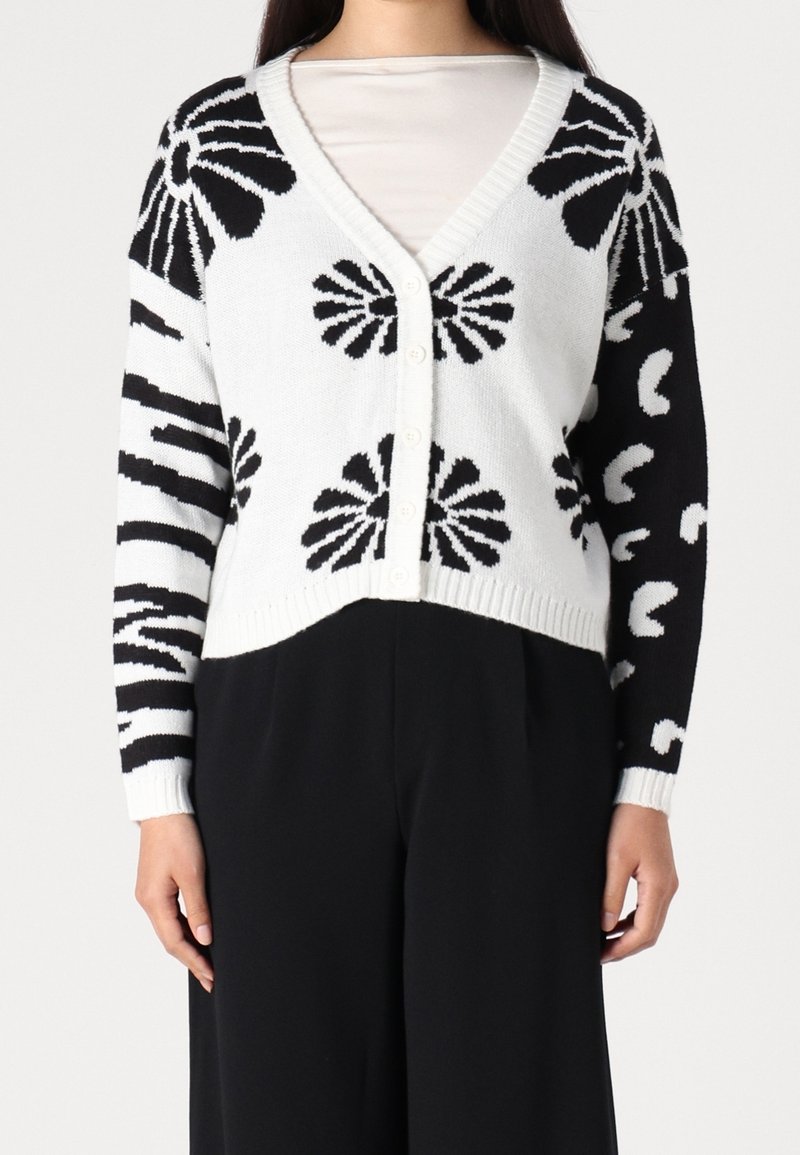 Cardigan blanc à boutons avec des motifs floraux abstraits noirs et des rayures, porté sur un haut blanc et un pantalon noir.