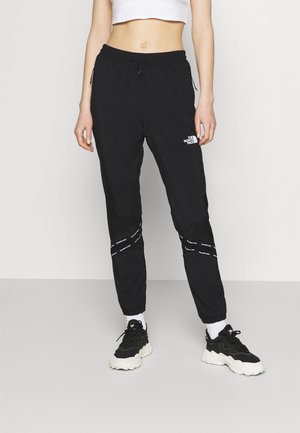 Personne portant un pantalon de jogging noir The North Face, un top court blanc, des chaussettes blanches et des baskets noir et blanc, debout devant un fond uni.