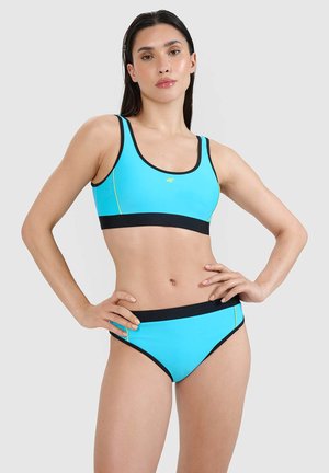 Kobieta w niebieskim bikini z czarną lamówką, stojąca z rękami na biodrach na jednolitym jasnoszarym tle.