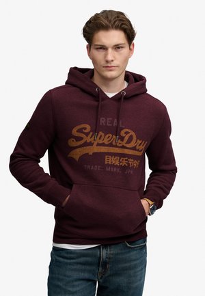 Joven con sudadera con capucha SuperDry marrón con las manos en los bolsillos delanteros y vaqueros azules, de pie frente a un fondo liso.
