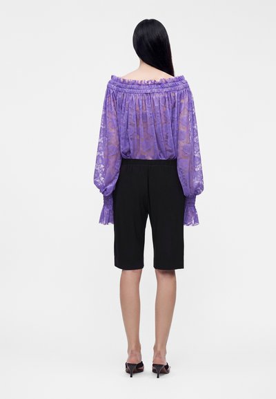 Femme aux longs cheveux noirs portant un chemisier violet transparent à épaules dénudées et un short noir au niveau des genoux avec des sandales à talons noirs, vue de dos.