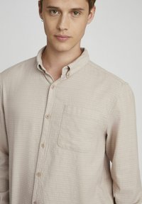 Chemise à boutons légère beige avec un col et une poche poitrine unique, présentant un tissu texturé et des boutons avant visibles.