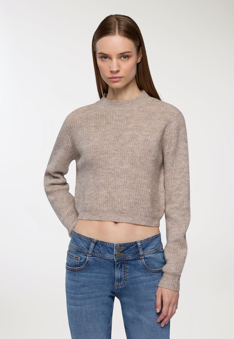 Kobieta z prostymi brązowymi włosami, ubrana w krótki beżowy sweter i niebieskie spodnie z wysokim stanem, stoi na tle jednolitego jasnego tła.