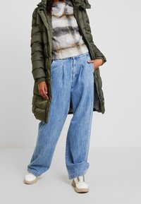 Olivenfarvet pufferjakke med hætte, grå ternet pelslignende overdel, lyseblå brede jeans og beige sneakers med hvide detaljer.