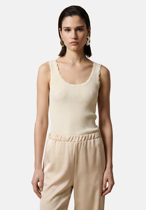 Donna con capelli corti e scuri che indossa grandi orecchini d'oro, canotta a coste color crema e pantaloni di raso beige su sfondo uniforme.