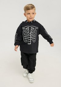 Felpa nera a maniche lunghe con stampa di scheletro bianca sulla parte anteriore e sulle maniche, abbinata a pantaloni cargo neri e sneakers bianche.