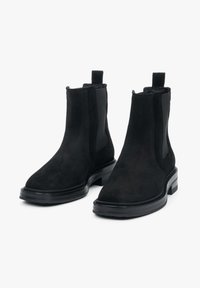 Para czarnych botków Chelsea zamszowych z elastycznymi panelami po bokach, pętelkami do ściągania i niskimi, kwadratowymi obcasami na białym tle.
