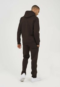 Brązowa bluza z kapturem i dopasowane spodnie wykonane z miękkiego materiału, wyposażone w kieszeń kangurką, regulowany kaptur i prążkowane mankiety. Białe sneakersy.